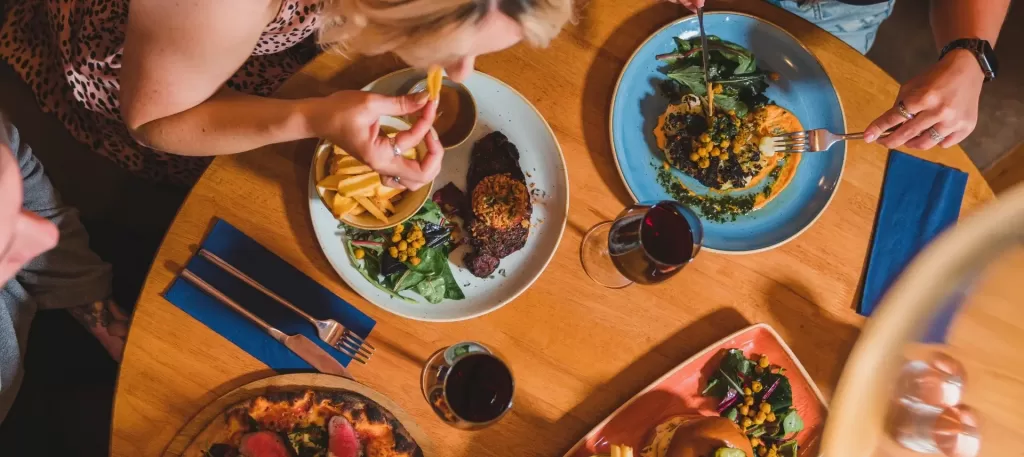 birds eye view of people eating food with multiple plates and glasses including steak, chips, chickpeas and blue napkins.