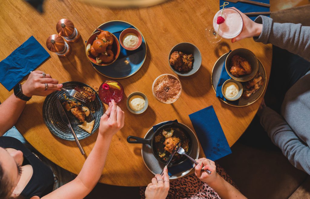 birds eye view of table with multiple plates of food and cocktails with three people sitting around.