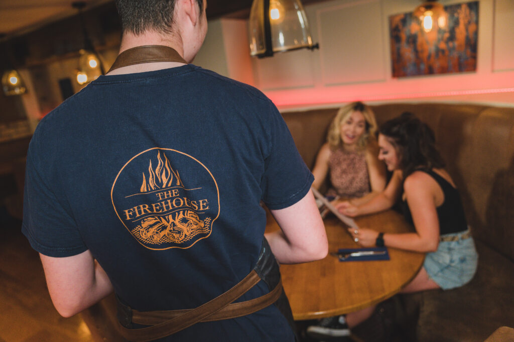 a man wearing a navy t-shirt with orange The Firehouse logo on the background walking to serve table. group diinner in enniskillen