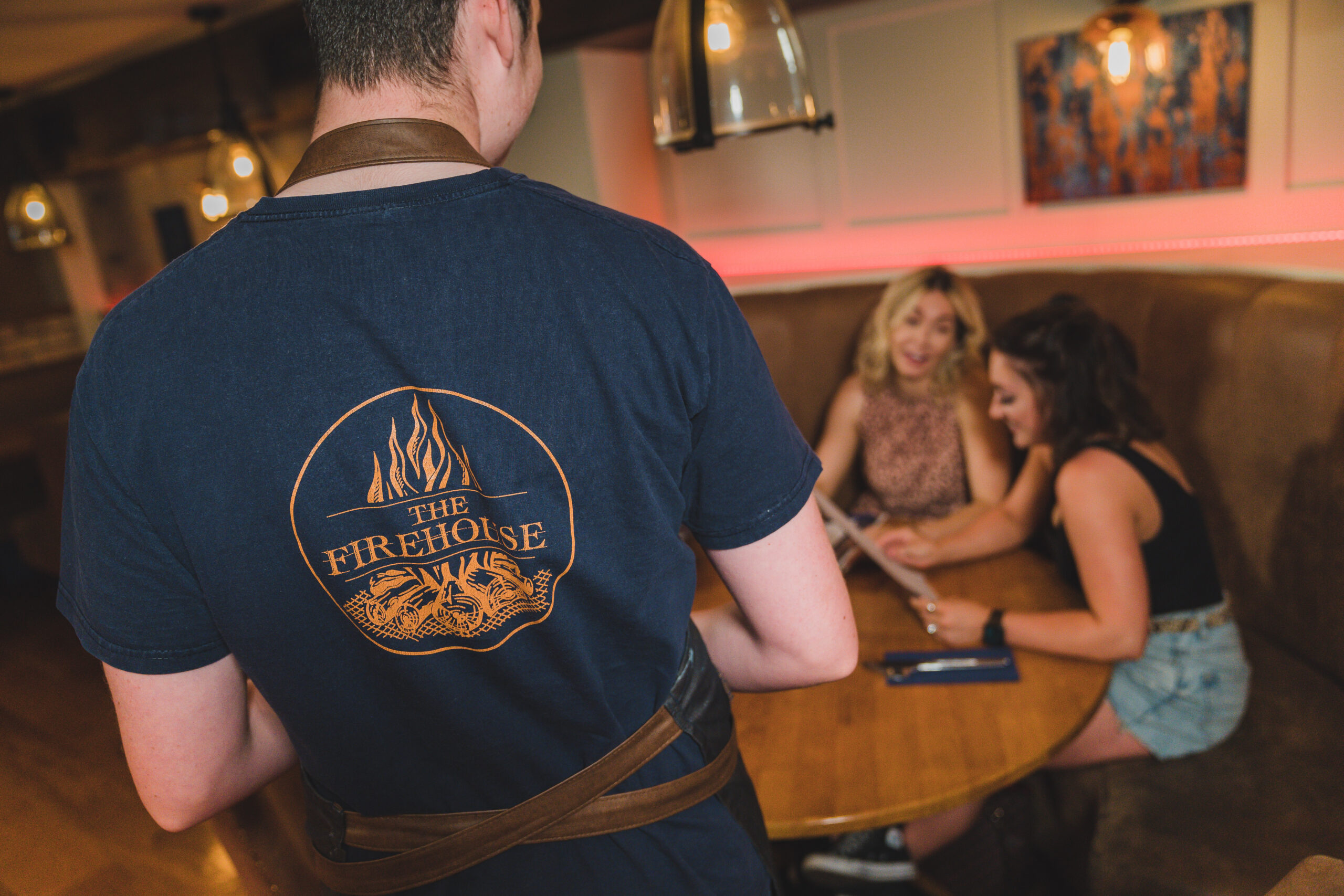 a man wearing a navy t-shirt with orange The Firehouse logo on the background walking to serve table. group diinner in enniskillen