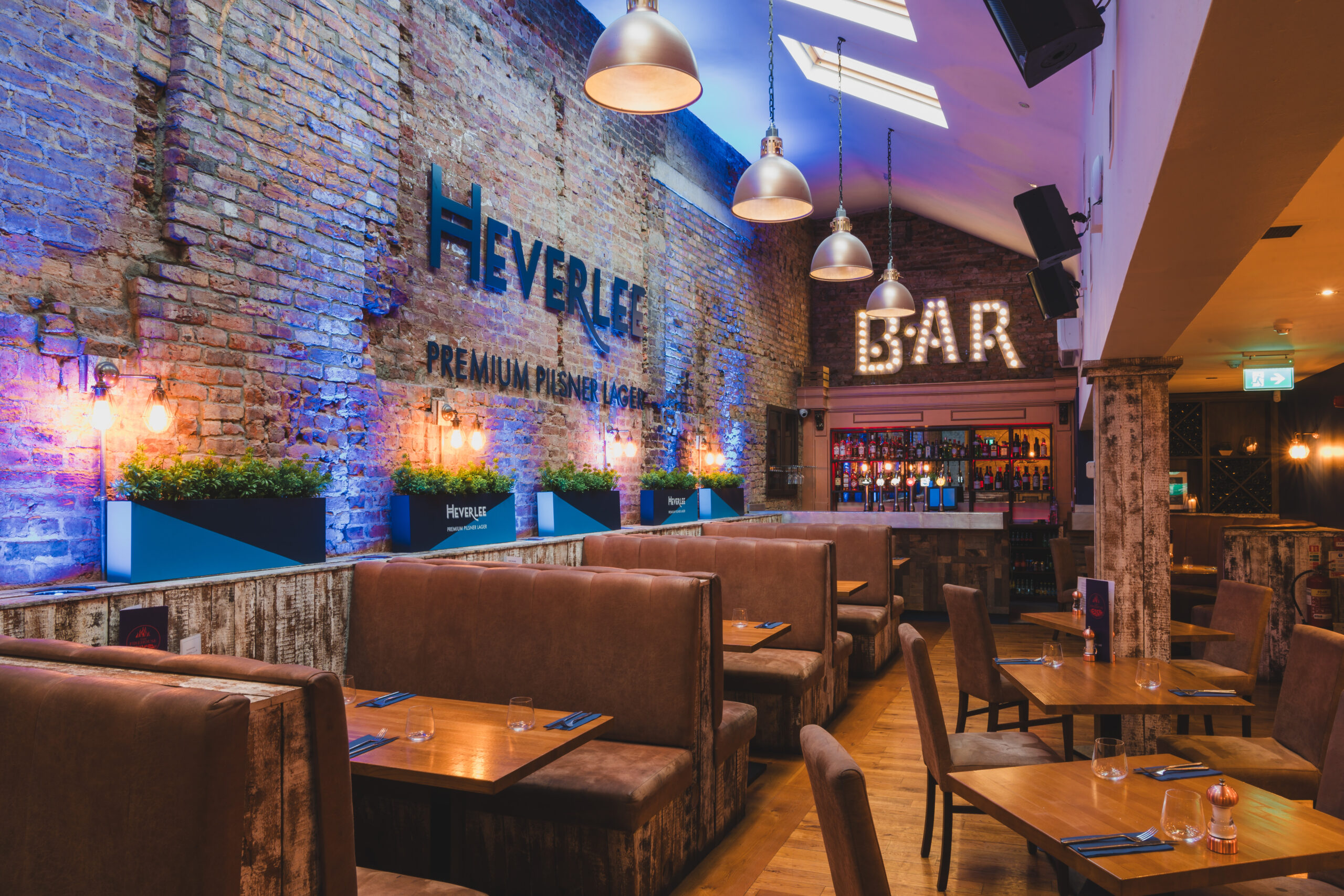 restaurant lit up with natural light in overhead windows. Brown booths and chairs with set tables and bar at the back of the room.